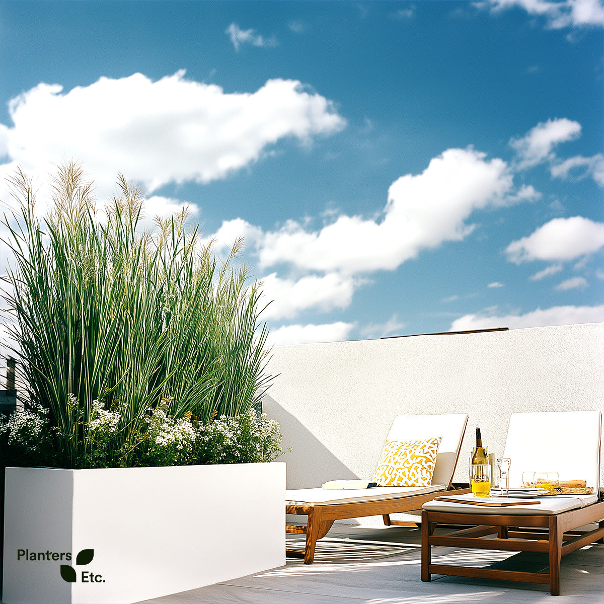 Rooftop patio with two wooden loungers and a sleek white rectangular planter filled with citronella grass and white flowers, set against a bright blue sky with scattered clouds.