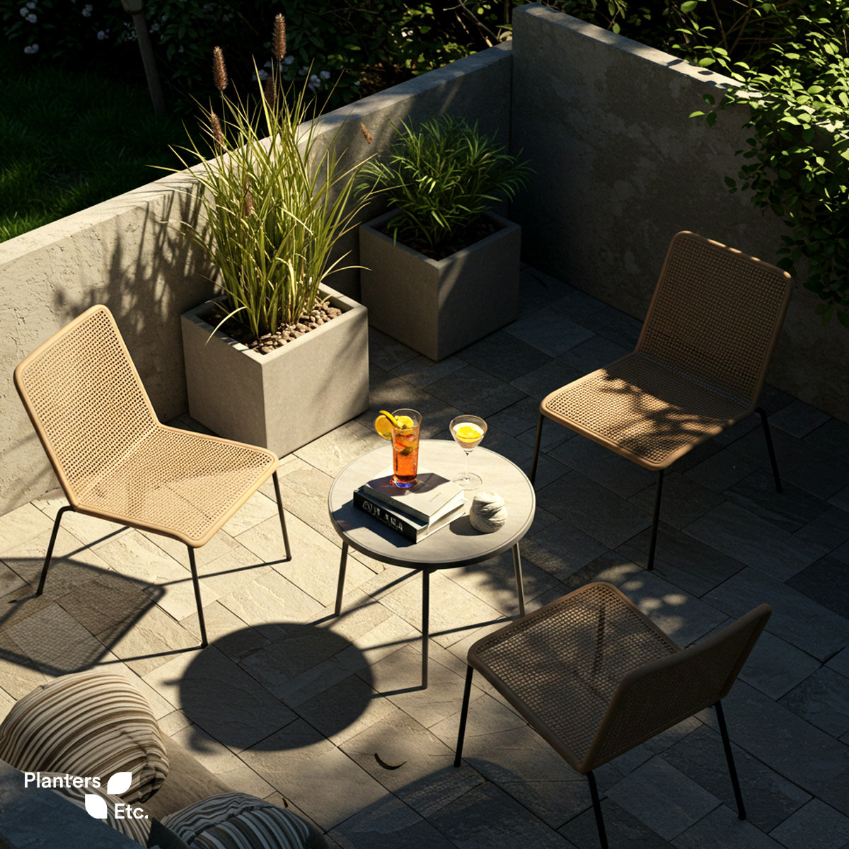 Modern stone patio with natural rattan chairs and a round metal table, surrounded by square planters filled with lemongrass and rosemary, basking in warm afternoon light.
