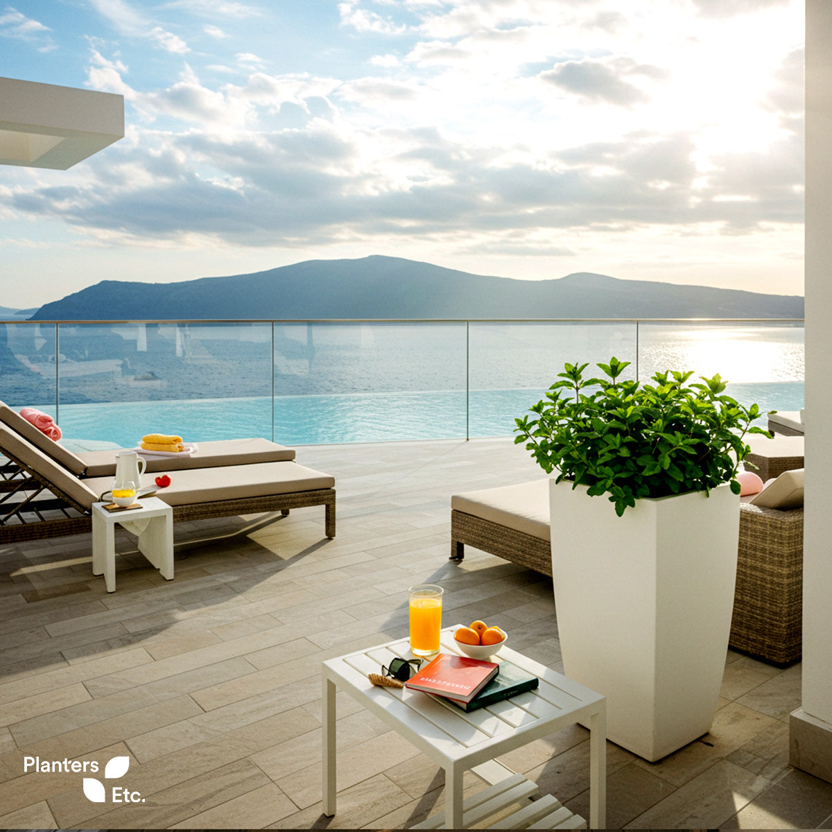 Modern rooftop terrace with ocean views featuring sun loungers, a white tapered planter filled with mint, and a small table set with juice, fruit, and books under soft morning light.